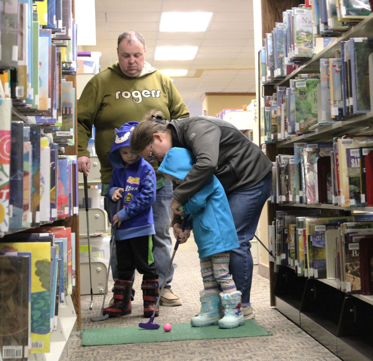 Woman helps small child make a putt at the indoor mini-golf event as man and older child watch