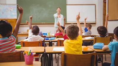 Kids raising their hands in a classroom with a teacher at the front of the classroom and a chalkboard behind them.